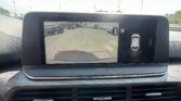 Rear-view camera display on a car dashboard showing a parking lot, with yellow and red guiding lines and the message 'Check surroundings for your safety.'