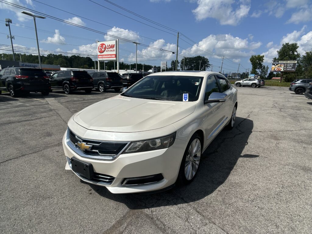 White Chevrolet sedan in foreground at a car dealership lot with several dark SUVs parked behind it, blue sky and power lines overhead, and a prominent E-Z Loan auto sales sign in the background.