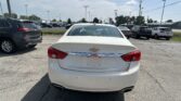 Rear view of a white Chevrolet sedan in a crowded car dealership parking lot, with other vehicles in the background and power lines overhead.