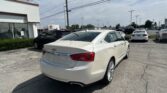 White Chevrolet sedan in the foreground at a car dealership lot with other cars and a white building to the left under a blue sky with power lines overhead.