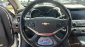 Inside view of a car cabin from the driver's seat showing a Chevrolet steering wheel, instrument cluster, and center console with a touchscreen/display, with a dealership lot visible through the windshield.